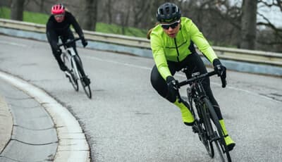 Two cyclists in professional gear racing around a curved road. The lead cyclist wears a neon yellow jacket.