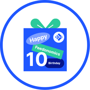 A celebratory graphic inside a blue circle featuring a blue gift box with a green bow. The box is decorated with the Feedonomics logo and stickers that say "Happy," "Feedonomics," "10," and "Birthday."