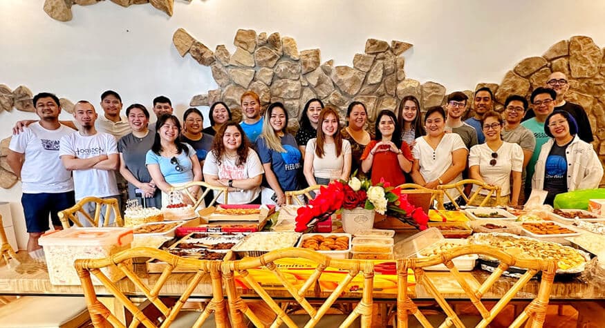 Large group of employees standing behind a long table filled with catered food and pizza.