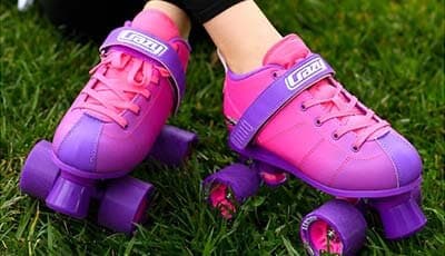 Pink and purple Crazy Skates roller skates worn by a person sitting on green grass.