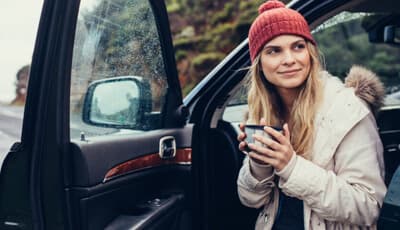 A smiling woman wearing a red beanie and a beige parka, holding a thermos while sitting in the open door of a car on a rainy day.