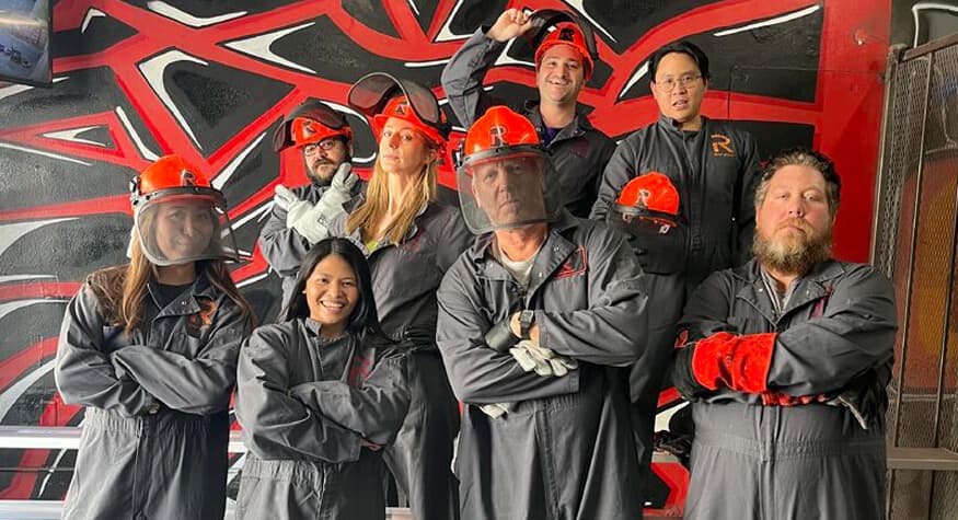 Team members in grey jumpsuits and orange safety helmets posing in front of a red and black mural.