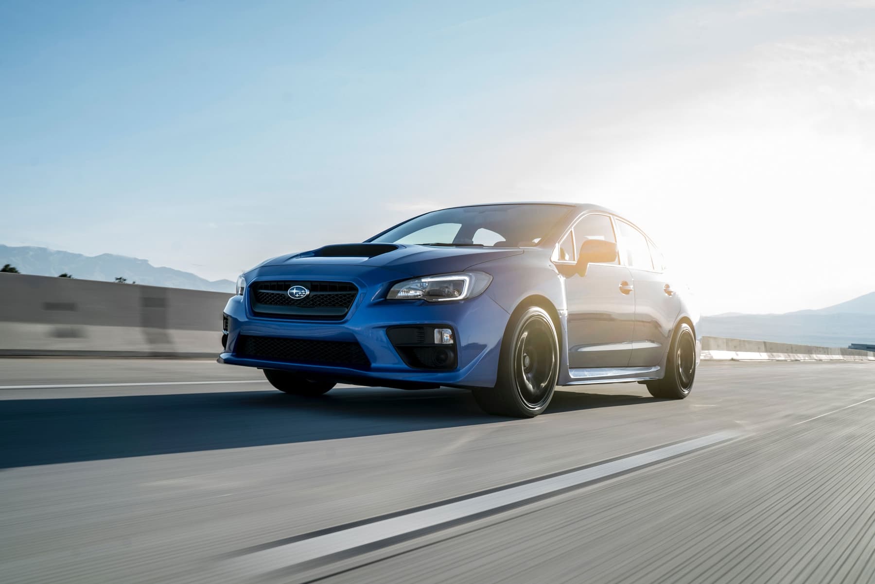 Dynamic, low-angle shot of a blue Subaru WRX driving at high speed on a highway during a bright, sunny day.