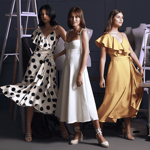 Three models posing in an atelier, wearing a black polka dot wrap dress, a white sundress, and a yellow ruffled wrap dress.