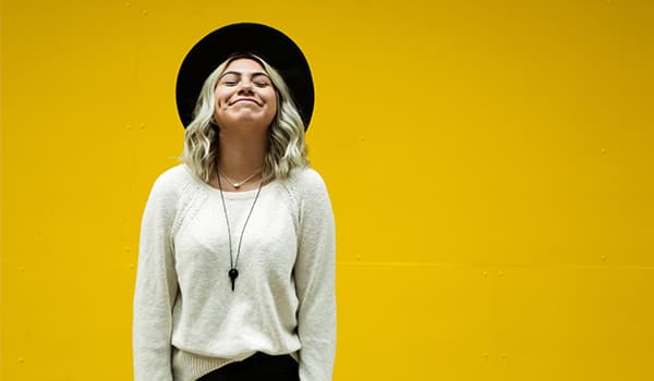 Woman in a white sweater and a black hat smiling upwards in front of a yellow backdrop