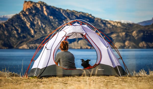 Woman and a dog sitting in a tent with a lake and mountain in the background